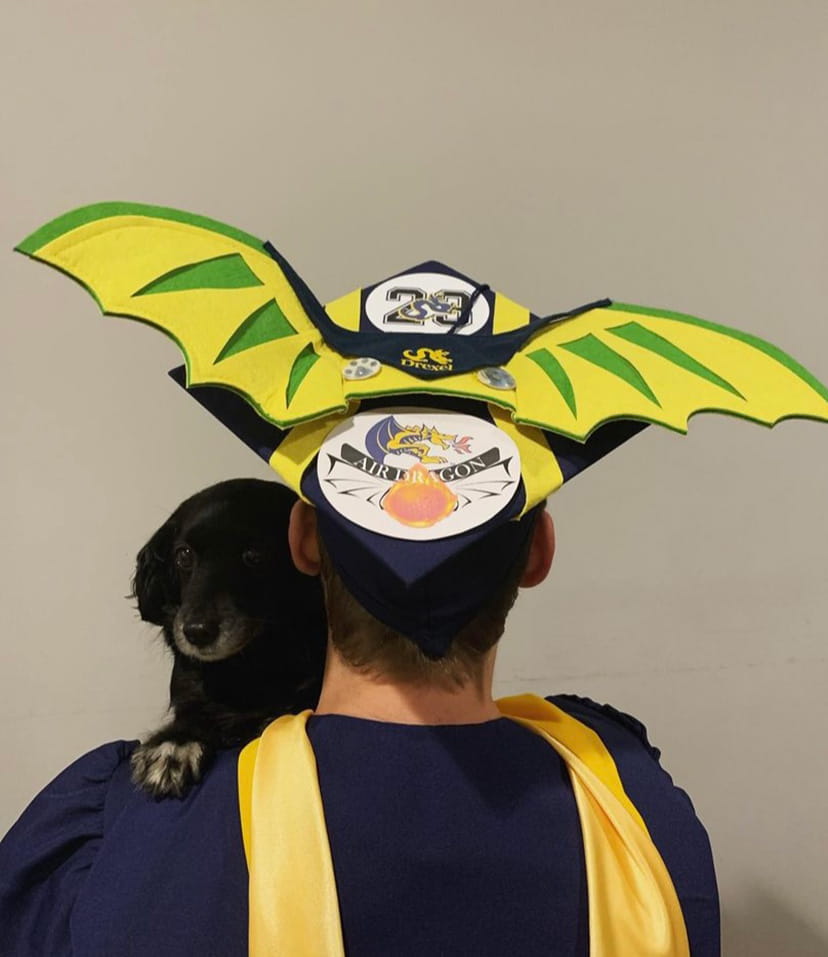 A graduate wears a commencement cap and gown while holding a dog. The commencement cap has yellow and green wings. 