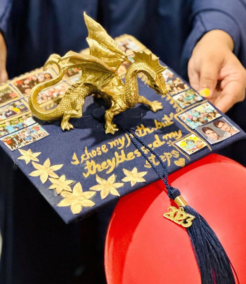 A 2023 commencement cap with decorated gold stars and photos and a 3d golden dragon figurine on top. 