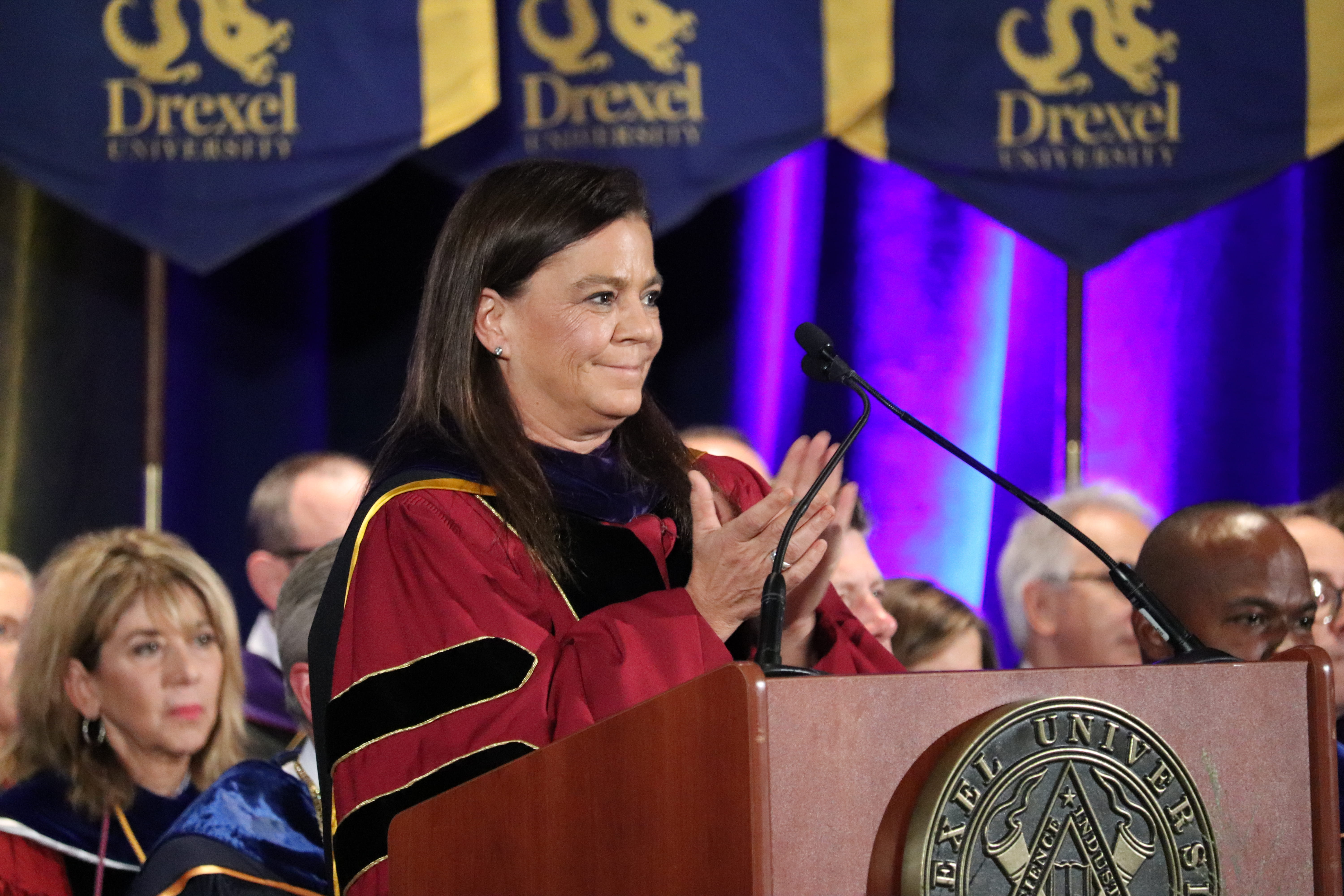 A woman clapping at a podium in Commencement gear.