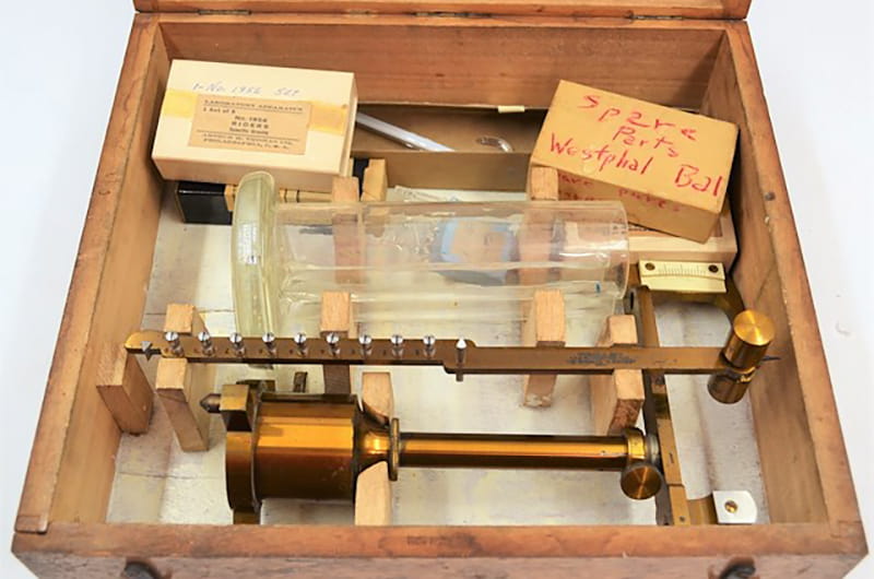 Wooden box containing a Westphal Specific Gravity Balance, a scientific instrument used for measuring specific gravity (ratio of the density of an object or substance to the density of standard substances like water or air). Pieces include brass stand and balance arm, a cylindrical glass vessel, a glass plummet, a curved glass thermometer (with mercury inside), and numerous metal scale riders and hooks.