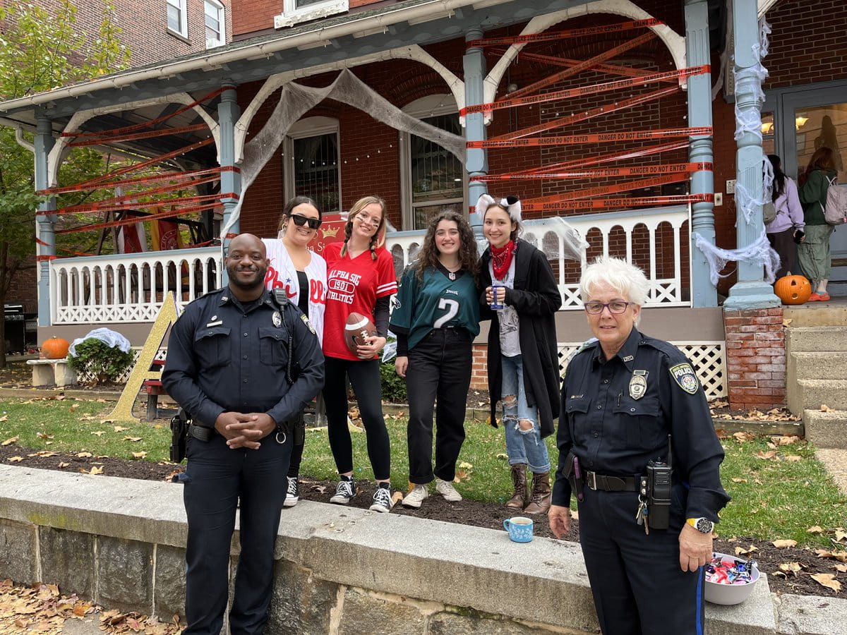 Two police officers and four female students in Halloween costumes standing outside a house decorated for Halloween.