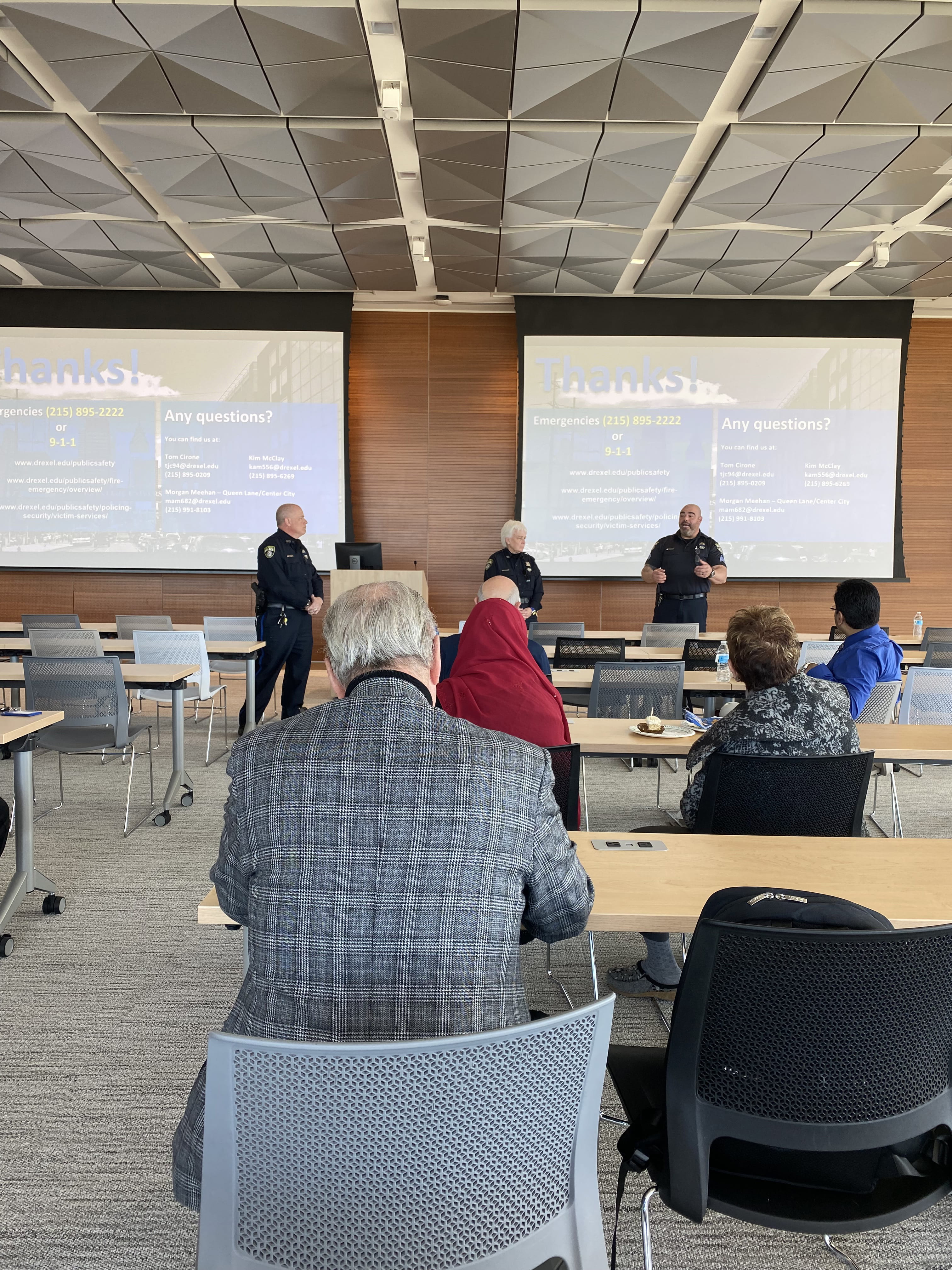 People sit in chairs and watch two police officers in front of two pull-down projection screens during a session.