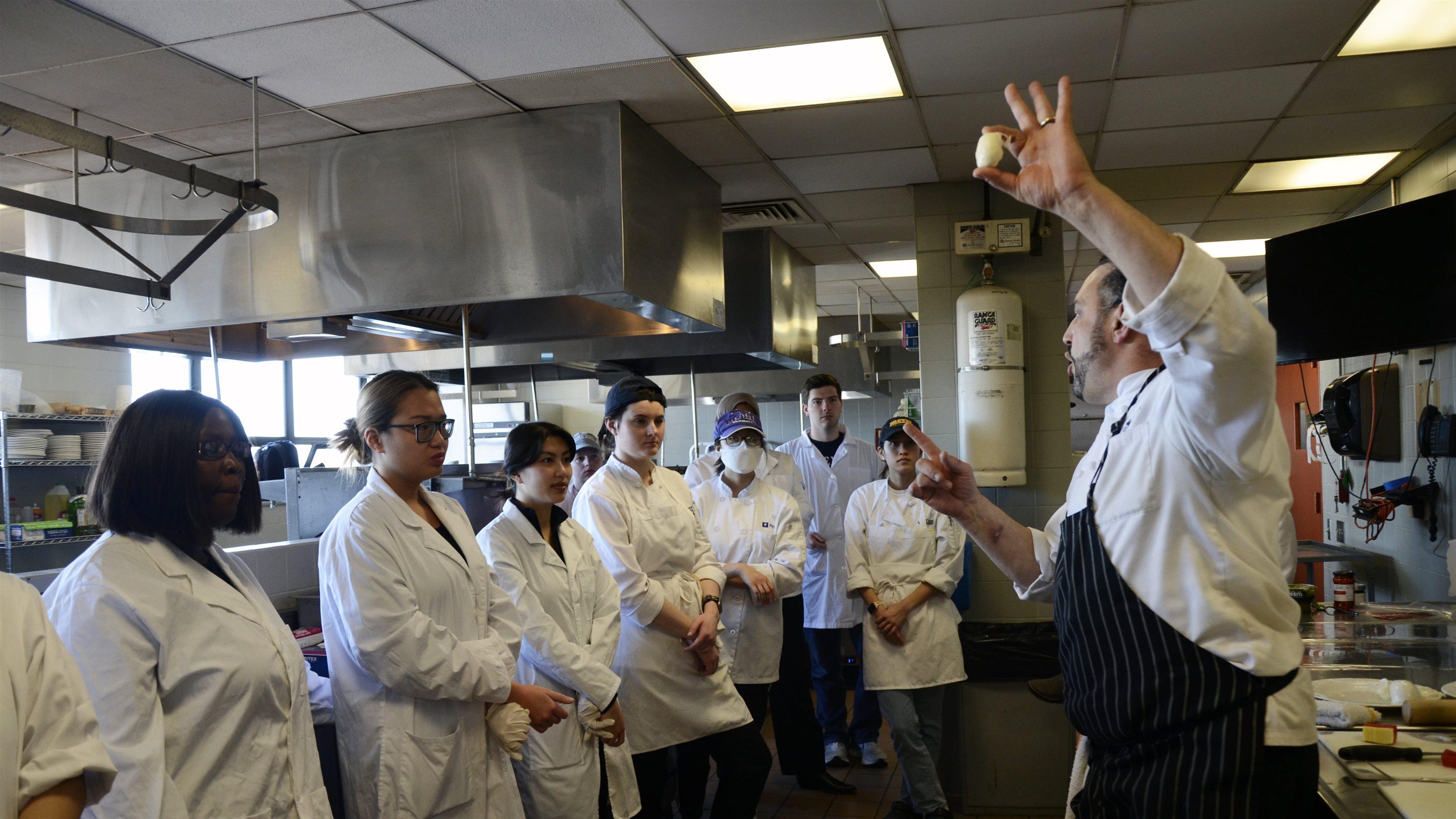 A man in a chef coat and black apron holds a piece of mozzarella up as a group of students in white coats observe.