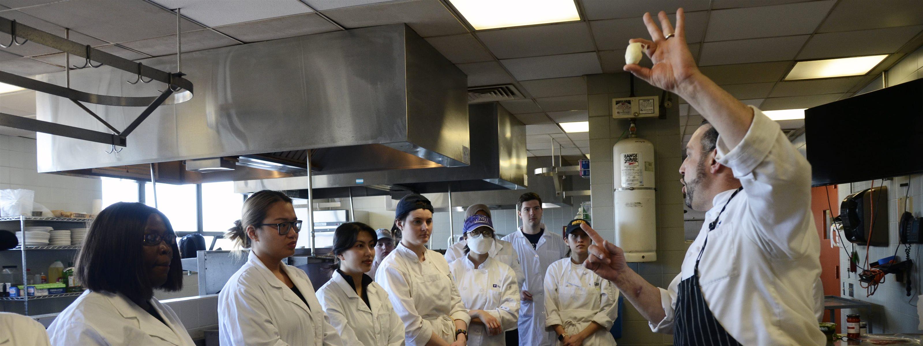A man in a chef coat and black apron holds a piece of mozzarella up as a group of students in white coats observe.