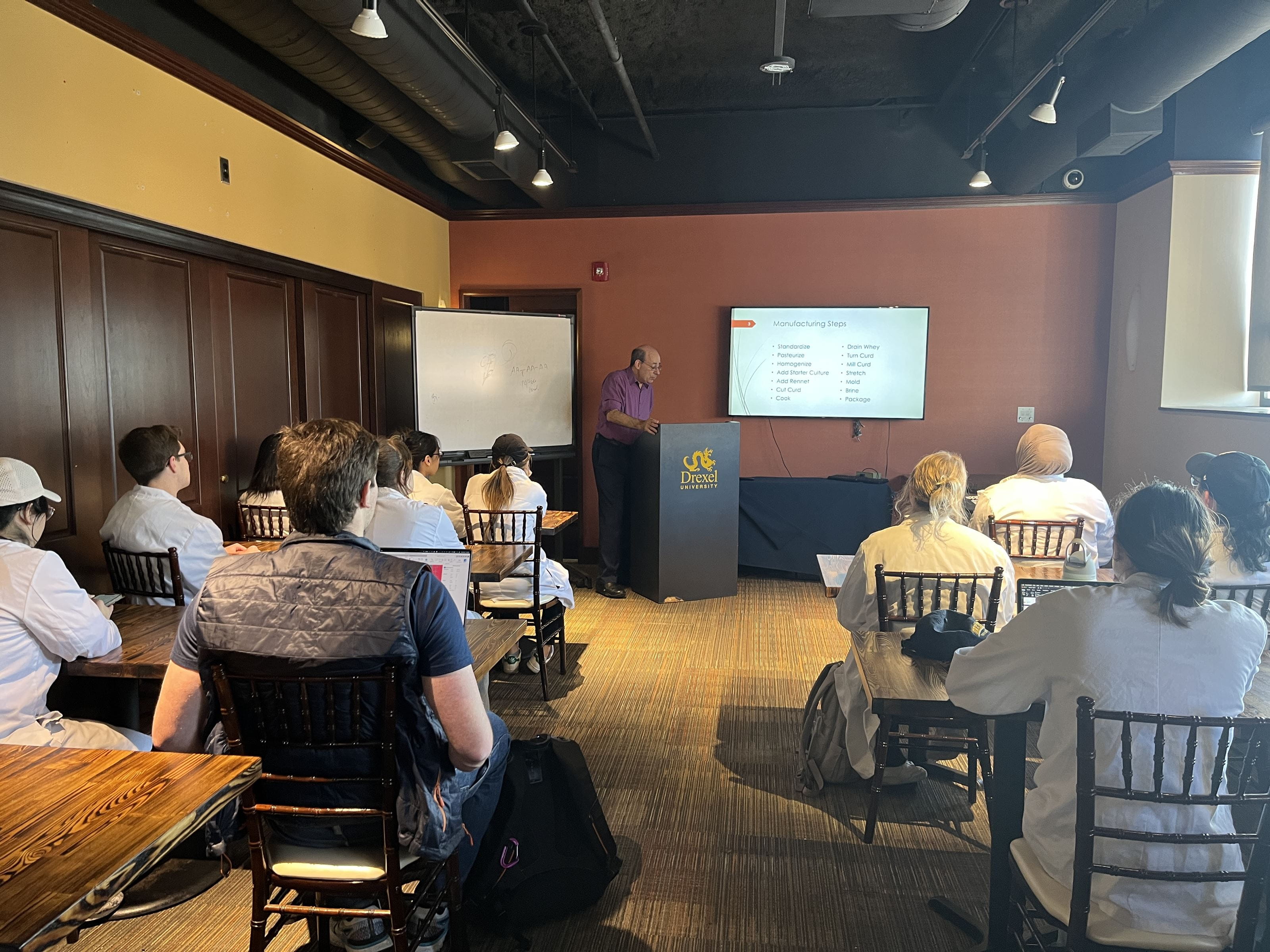 Students sit in a room looking at a powerpoint presentation presented by a man at a podium.