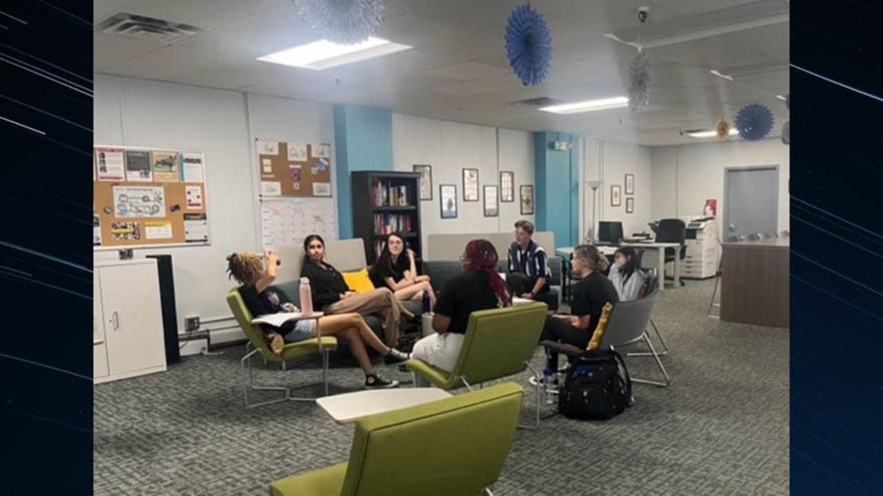 A group of students sitting on green couches and chairs in a room. 