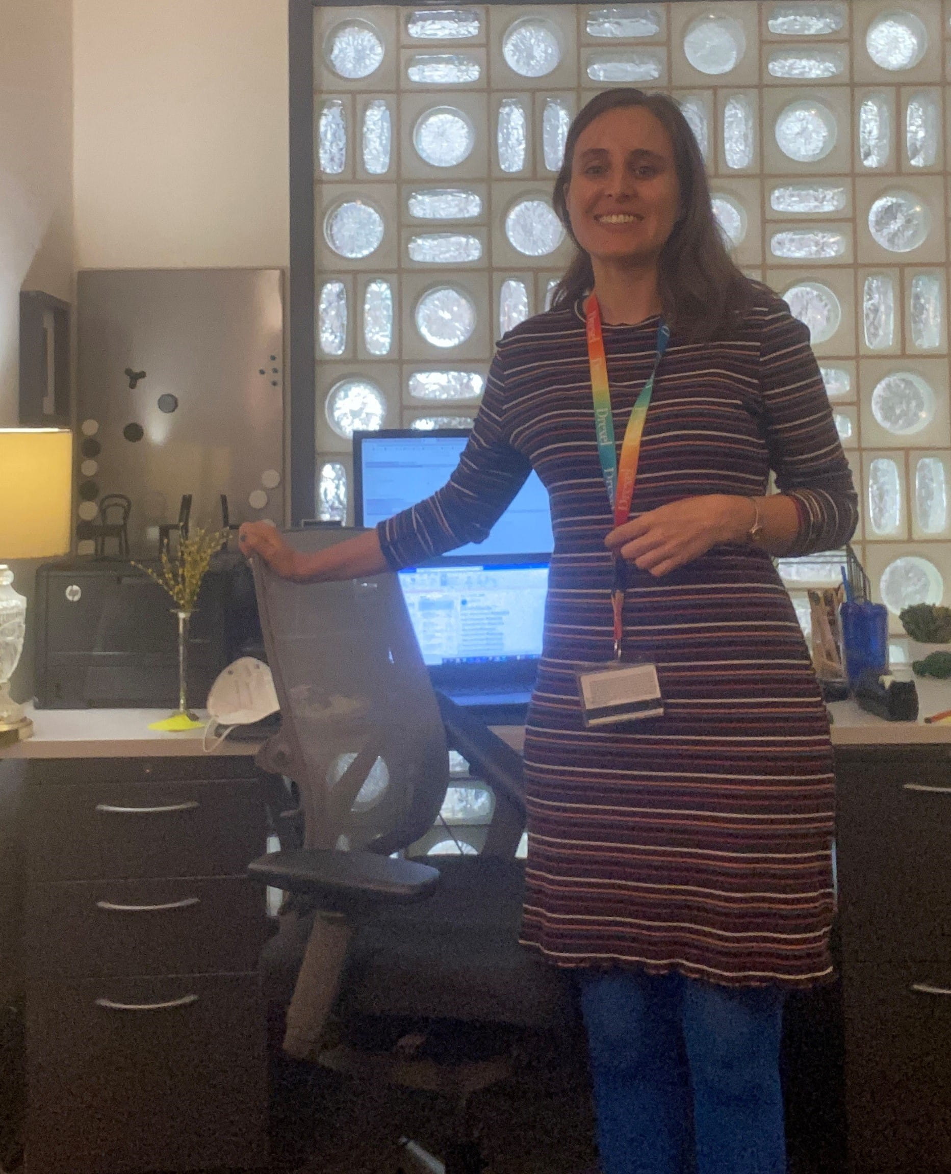 A woman in a striped dress stands next to the chair of her office setup.