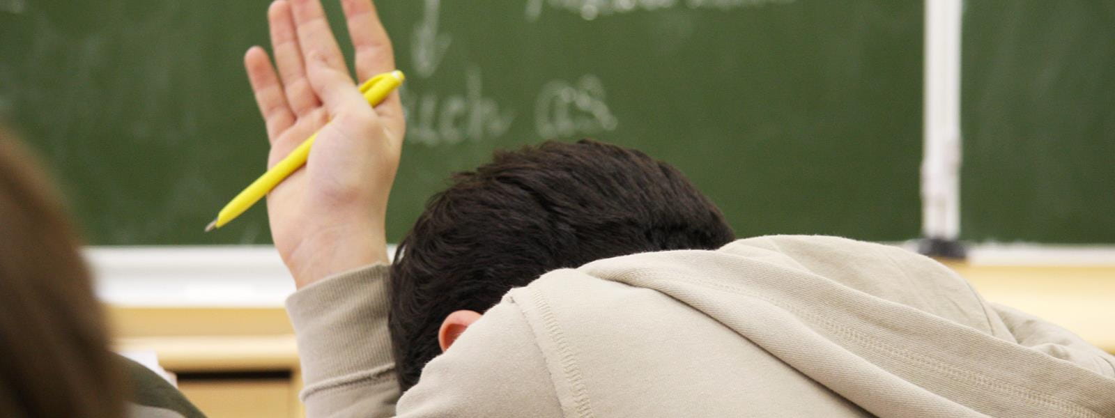 Young boy with his head down raises his hand with a pen.