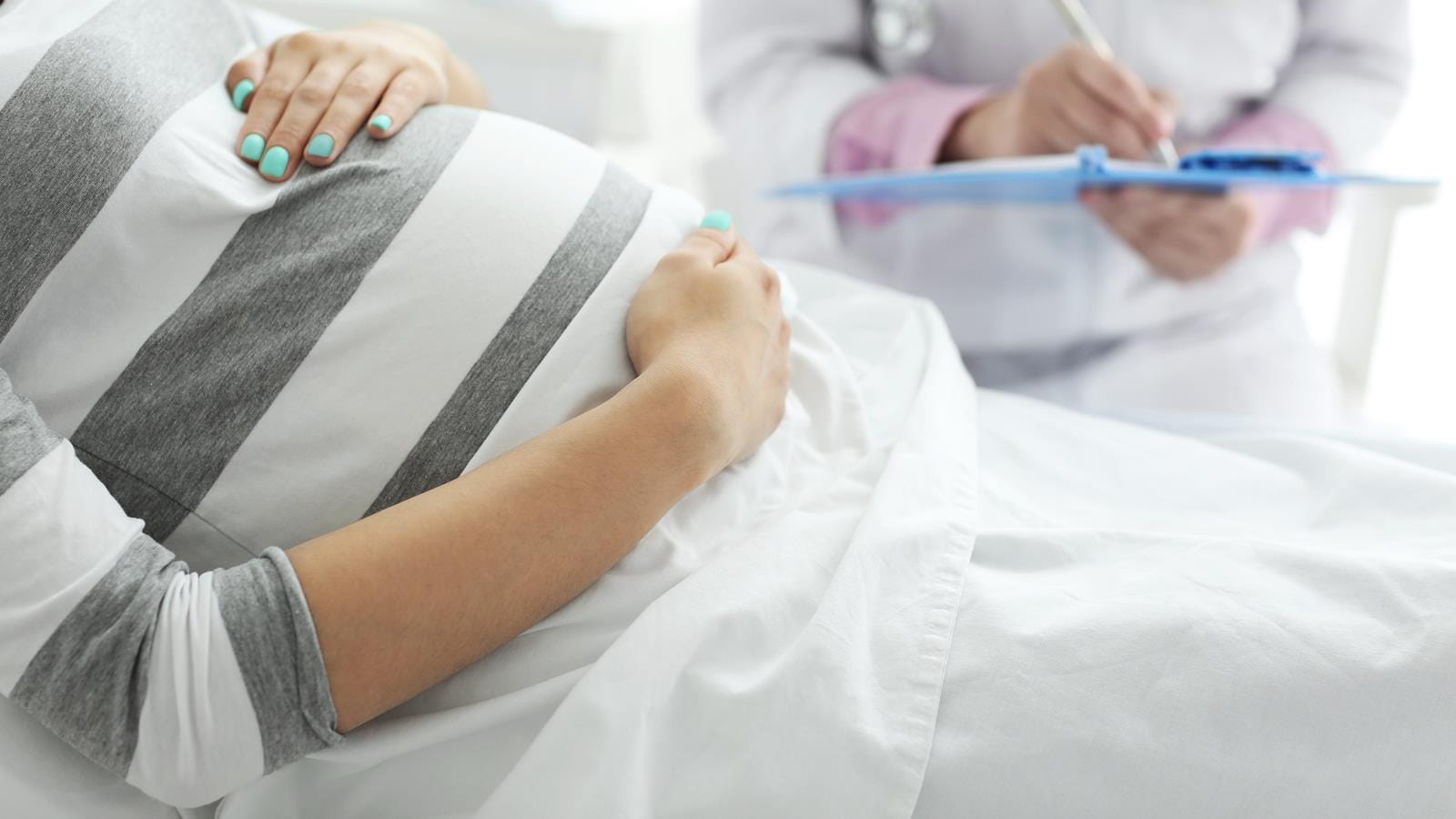 Pregnant person laying down holding their belly while a doctor stands with a clip board and white coat in the background.