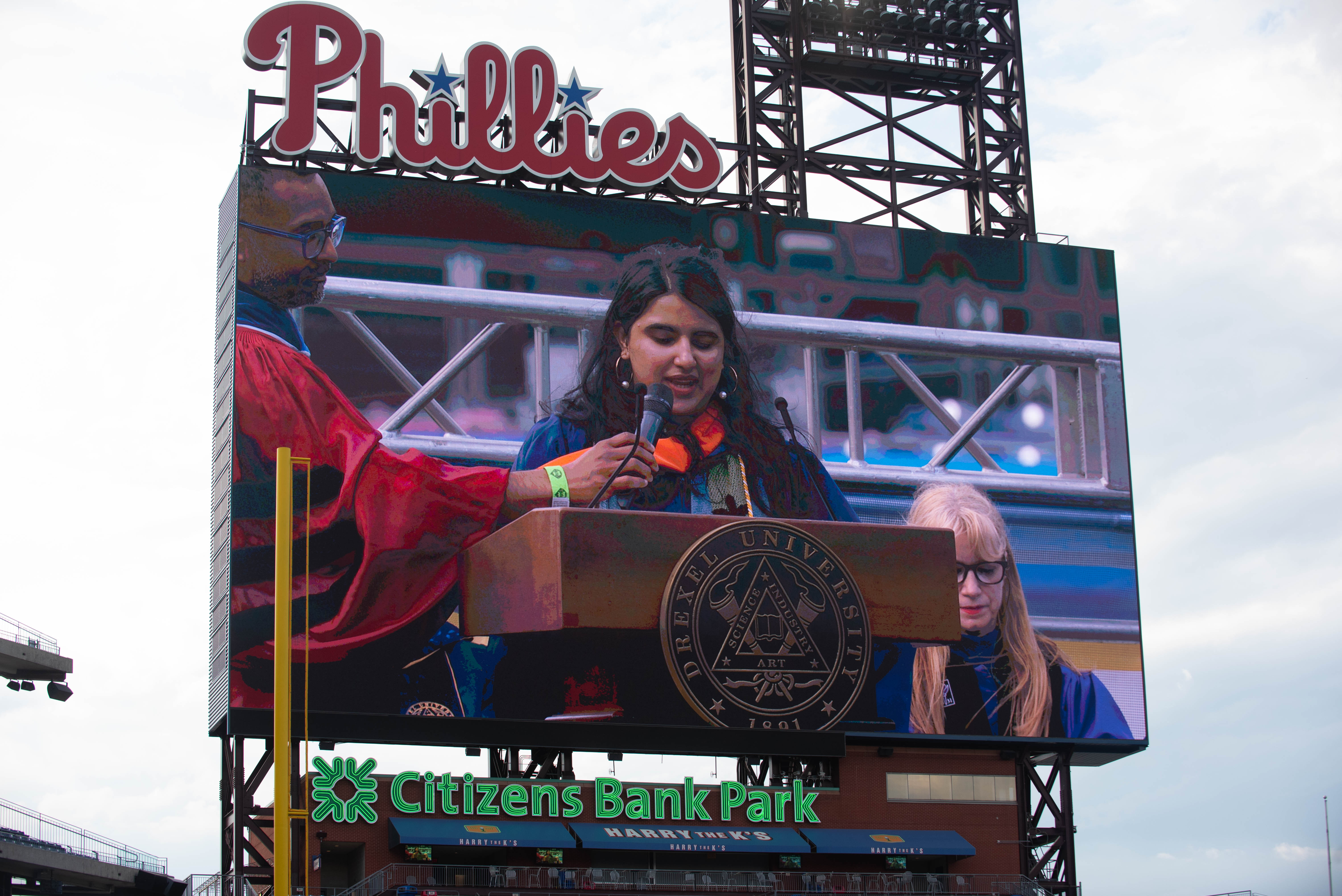 A screen showing Cimi Patani speaking at the podium at Drexel University's 2024 Commencement ceremony.