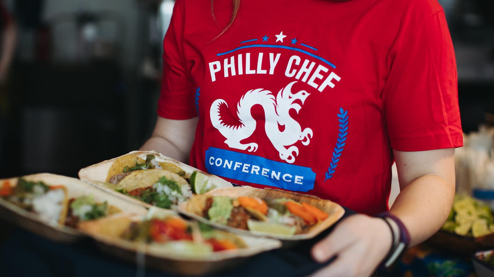 A woman in a red shirt with a Philly Chef Conference logo holding a tray with food-filled, small tasting plates