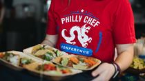 A woman in a red shirt with a Philly Chef Conference logo holding a tray with food-filled, small tasting plates