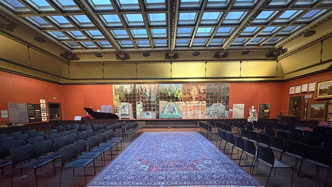 The view from the entrance of the A. J. Picture Gallery showing a tiled mural on a wall in a room full of chairs and art on other walls.