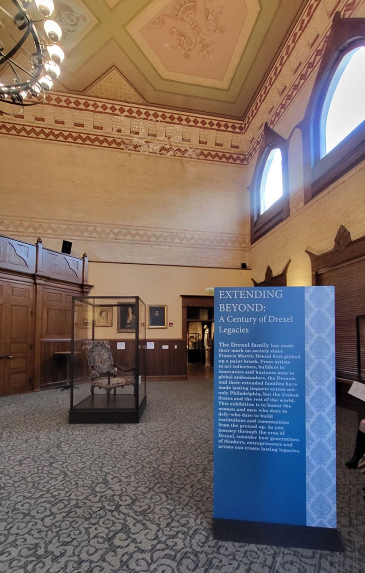 The right side of the room showcases the standing sign introducing the exhibition, with an early 18th century chair displayed behind it, also donated by Philip Syng Physick Fell. On the wall are portraits (from left to right) of Pauline Munn Doyle, her mother Mary Astor Paul Allez, and Alice Troth Drexel. Photo by Gina Monahan.