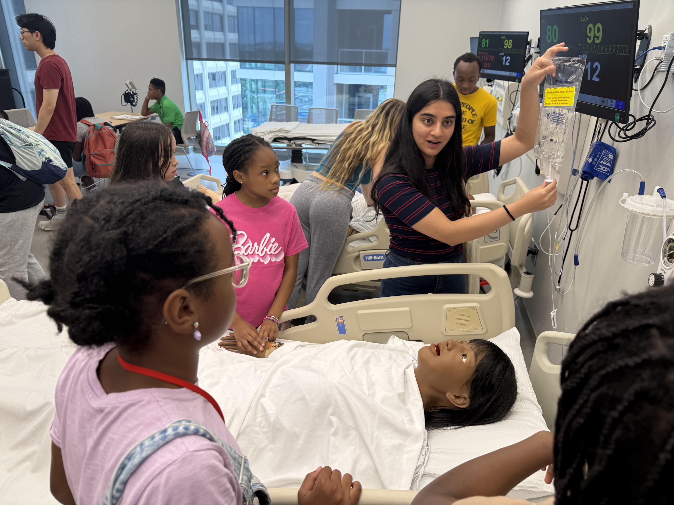 Middle school students around a hospital bed with a mannequin and a college student gesturing to the object.