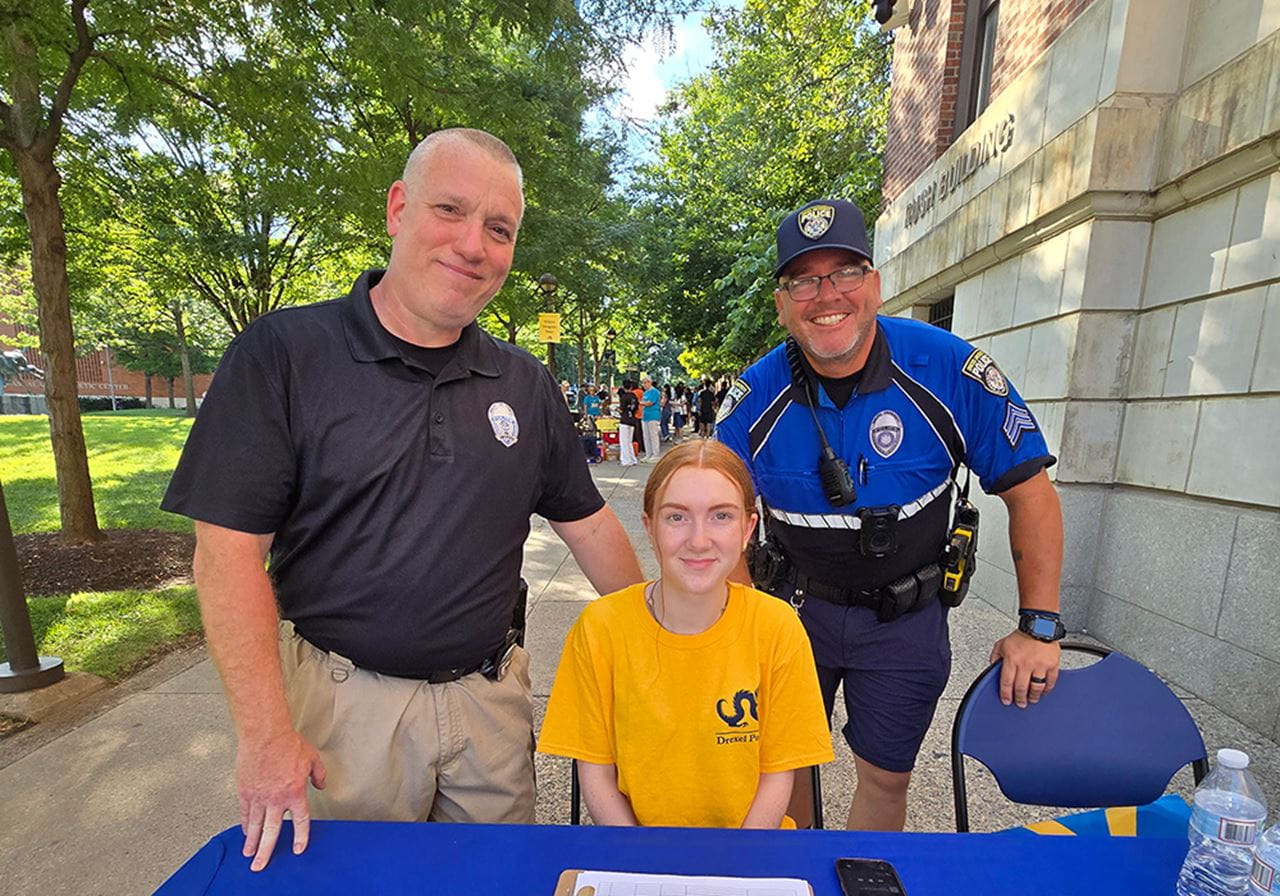 A female student in a gold Drexel shirt sits at a table with two male Drexel Public Safety officers standing behind her.