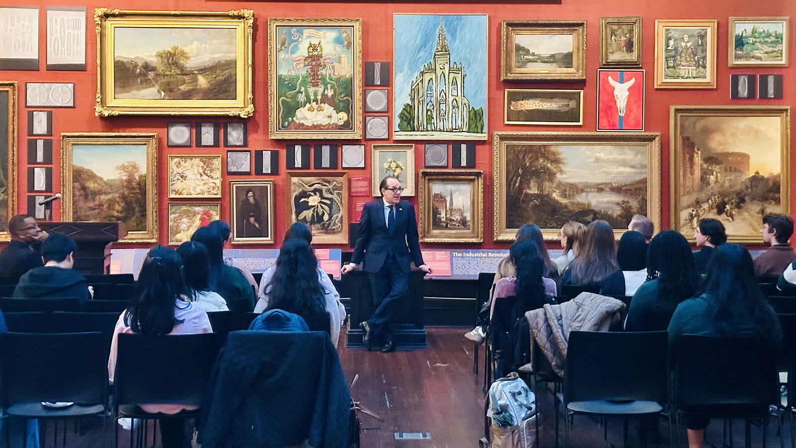 Antonio Merlo stands in front of seated students in a room with red wallpaper and lots of framed artwork behind him.