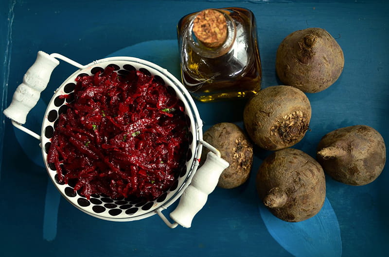 A bowl of red shredded beets, next to dirty whole beets on a blue table.