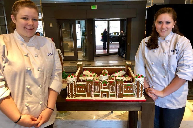 Melissa Martin, left, and Katelyn Comerford stand with the finished Eastern State Penitentiary gingerbread house at the Logan Hotel.
