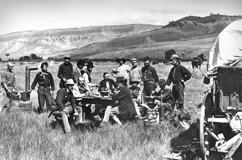 In this photograph of the Hayden Survey team, geologist and survey leader Ferdinand Hayden is seated cross-legged on the right side of the table and a bearded William Henry Jackson sits at the head of table facing camera. Although Jackson composed this photograph, a crew member would have taken the photo. Photo: "In camp at Red Buttes" (1870) by William Henry Jackson.