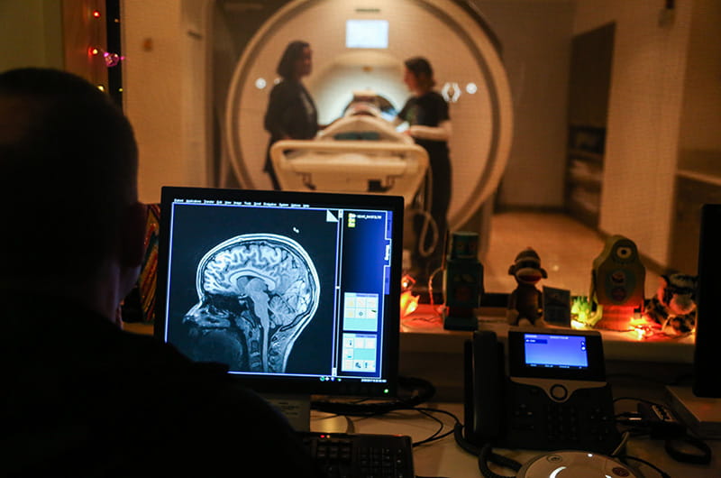 A person looking at a brain image on a monitor while someone else is helped into an MRI in the background.