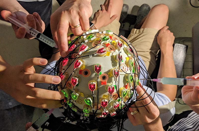 Researchers' hands preparing a test subject by dispensing conductive electrode gel into the electrodes in an EEG cap (top view of head).