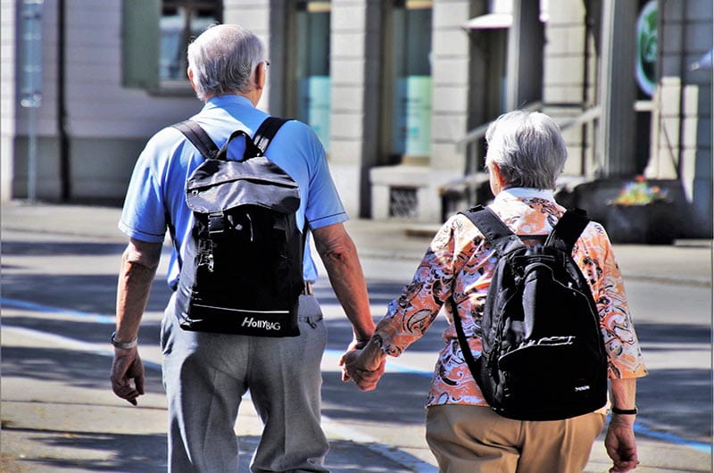 Older man and women holding hands and wearing backpacks. 