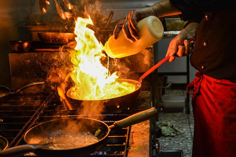 Chef pours oil into pan with flame in it. 