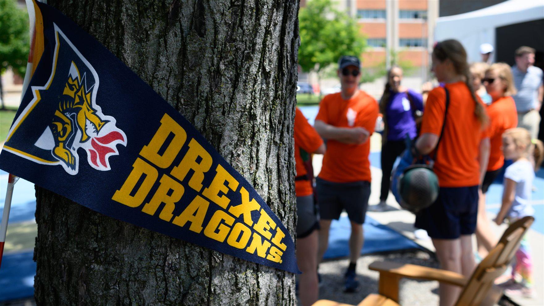 A &quot;Drexel Dragons&quot; flag pinned to a tree as participants of Employee Olympics gather in the background.