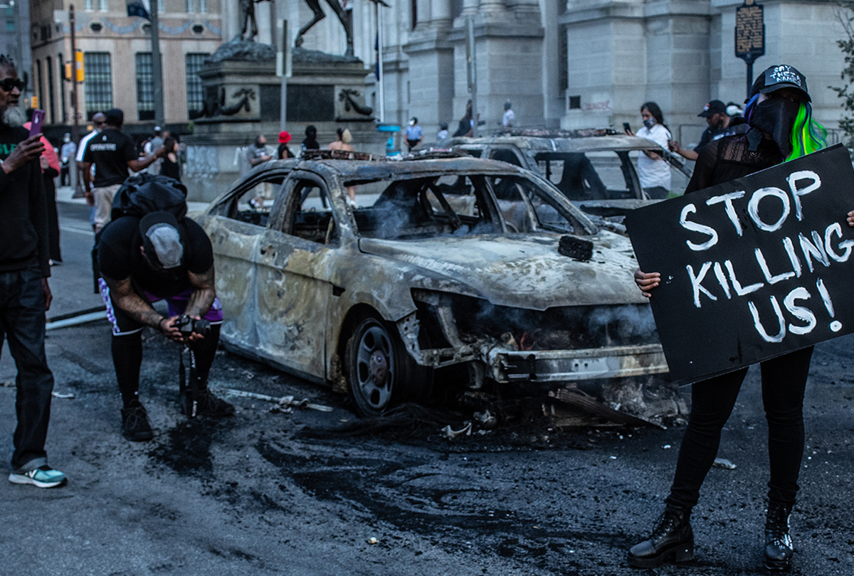 Two men stand in front of a burned car while a woman holds a sign saying "Stop killing us!"