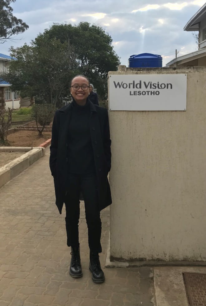 Woman in black coat stands in front of a sign reading "World Vision Lesotho."