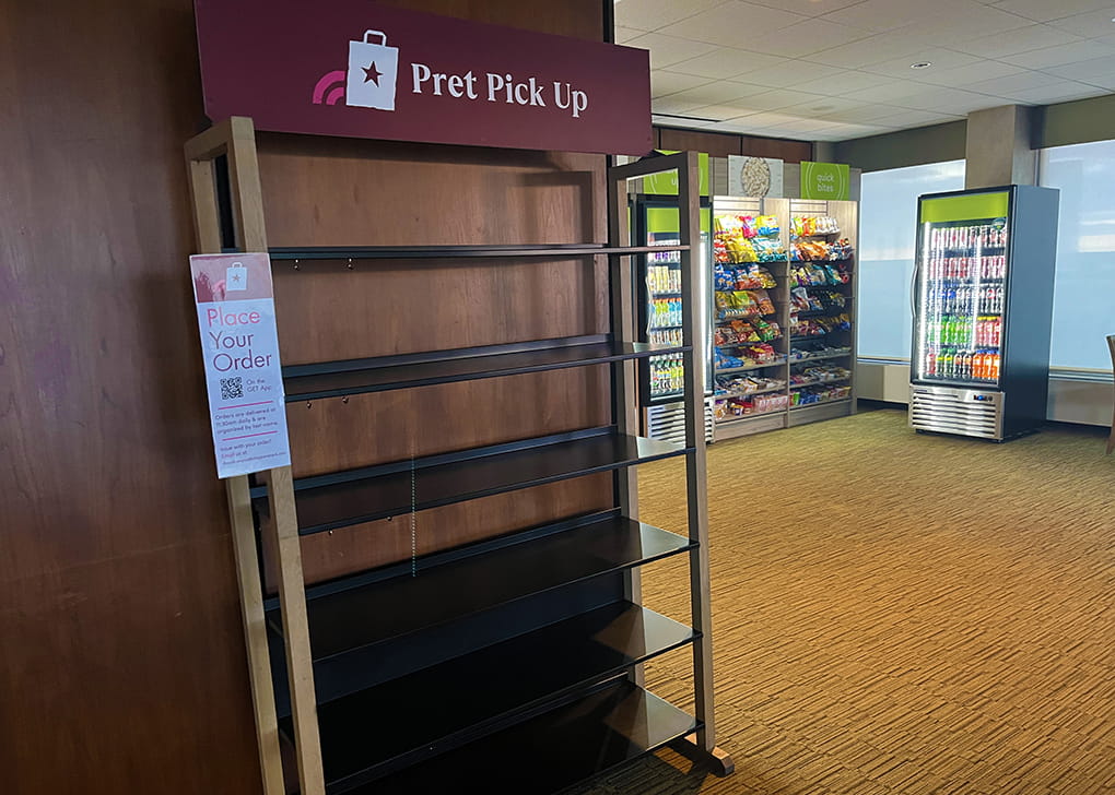 A shelving area with the words "Pret Pick Up" in front of a refrigerator of drinks and a wall of snacks to purchase.