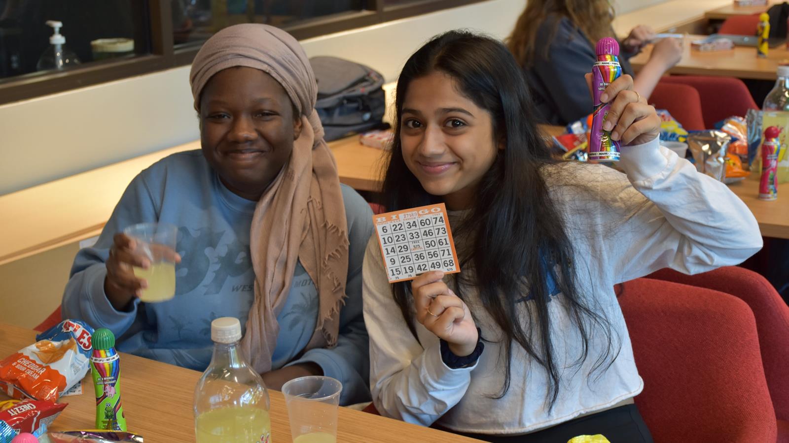 Two students smile at the camera. One is holding a cup of lemonade and the other is holding up a bingo card.