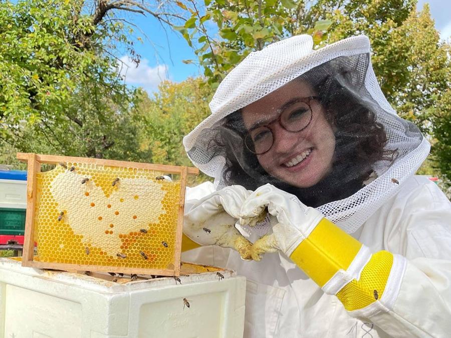 Smiling student in beekeeping gear, making a heart with her hands to mirror the heart shape of the honeycomb 