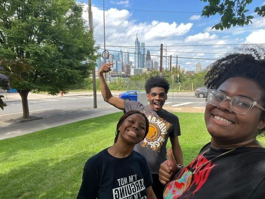 Three STAR Scholars taking a selfie in a park, one of them points at the Philly skyline