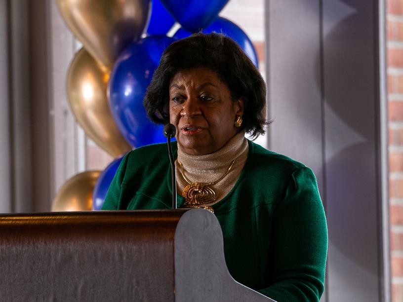 African American woman standing at a podium before gold and blue balloons