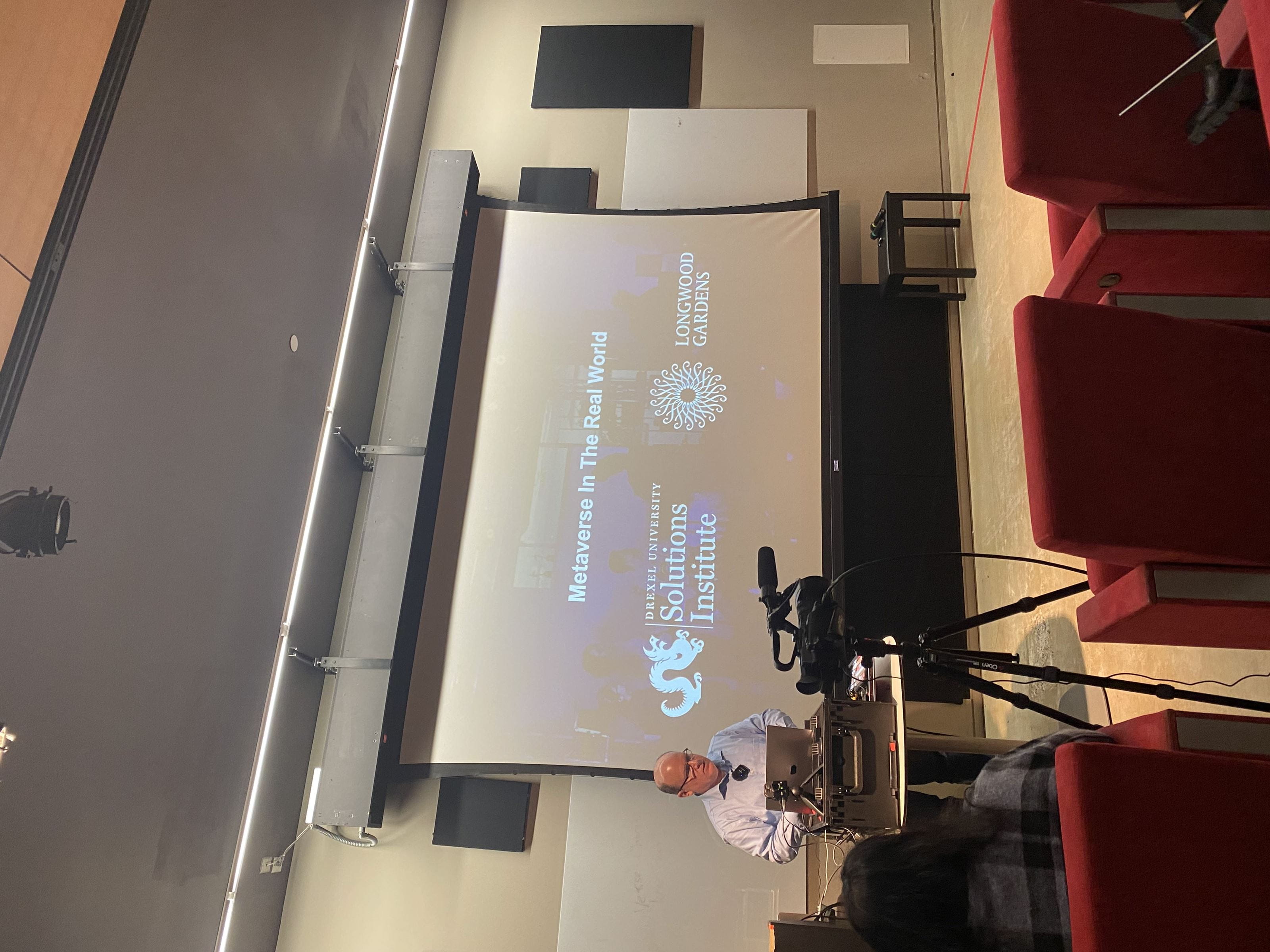 Associate Professor Nick Jushchyshyn standing in front a projection screen that reads "Metaverse in the Real World" with the Drexel University and Longwood Gardens logos underneath the text.