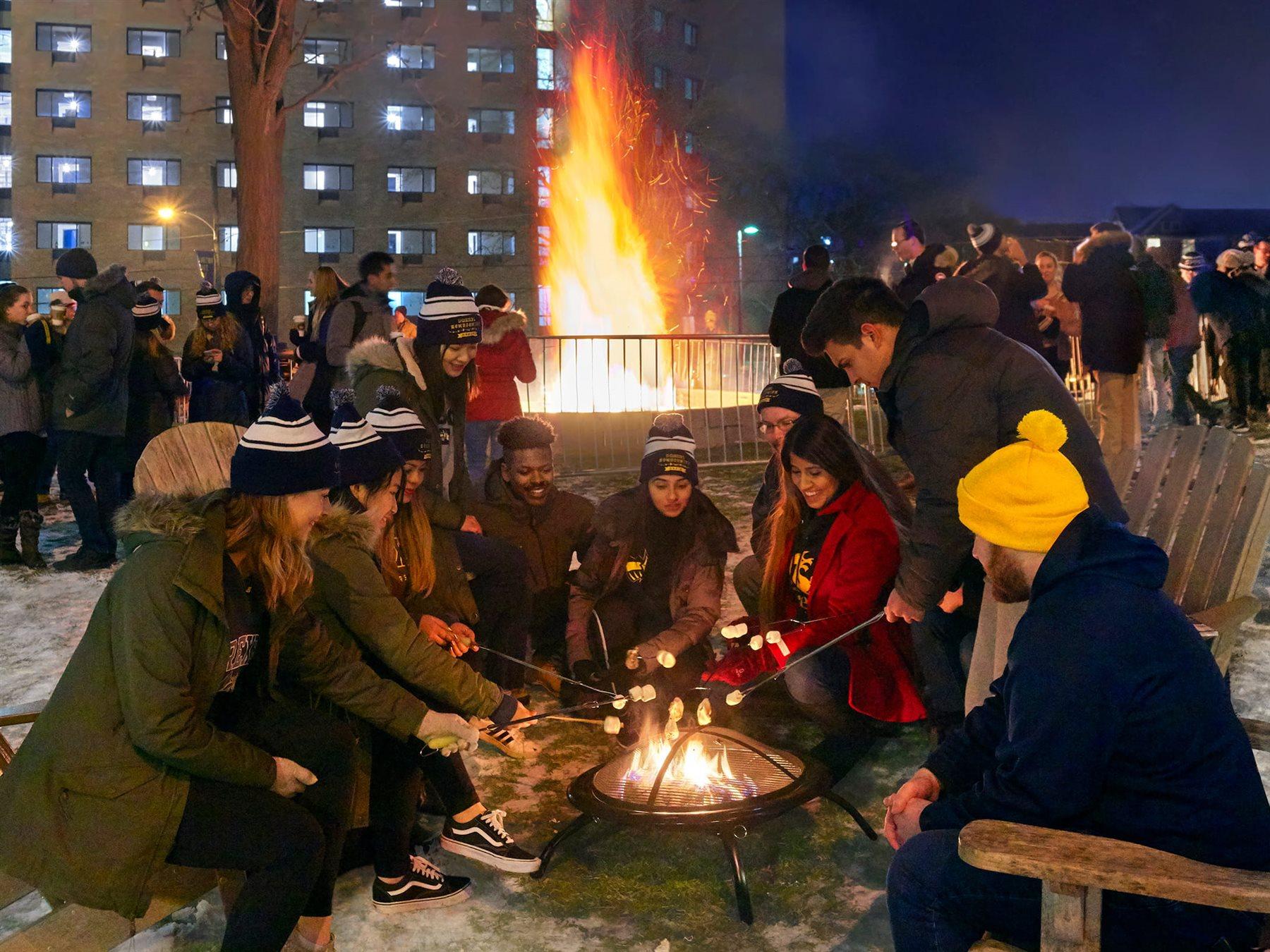Students sitting and roasting marshmallows over a fire, while other students stand and talk together on a snow-covered lawn
