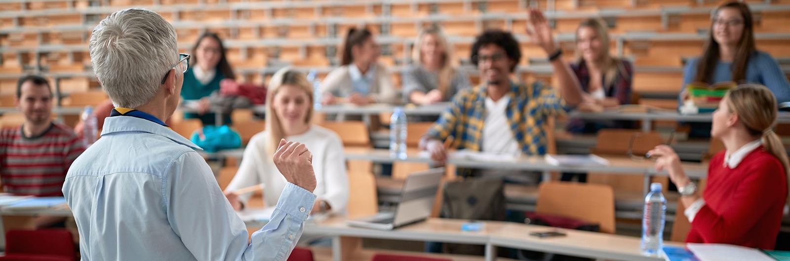 teacher stands in front of a lecture hall and students raise their hands