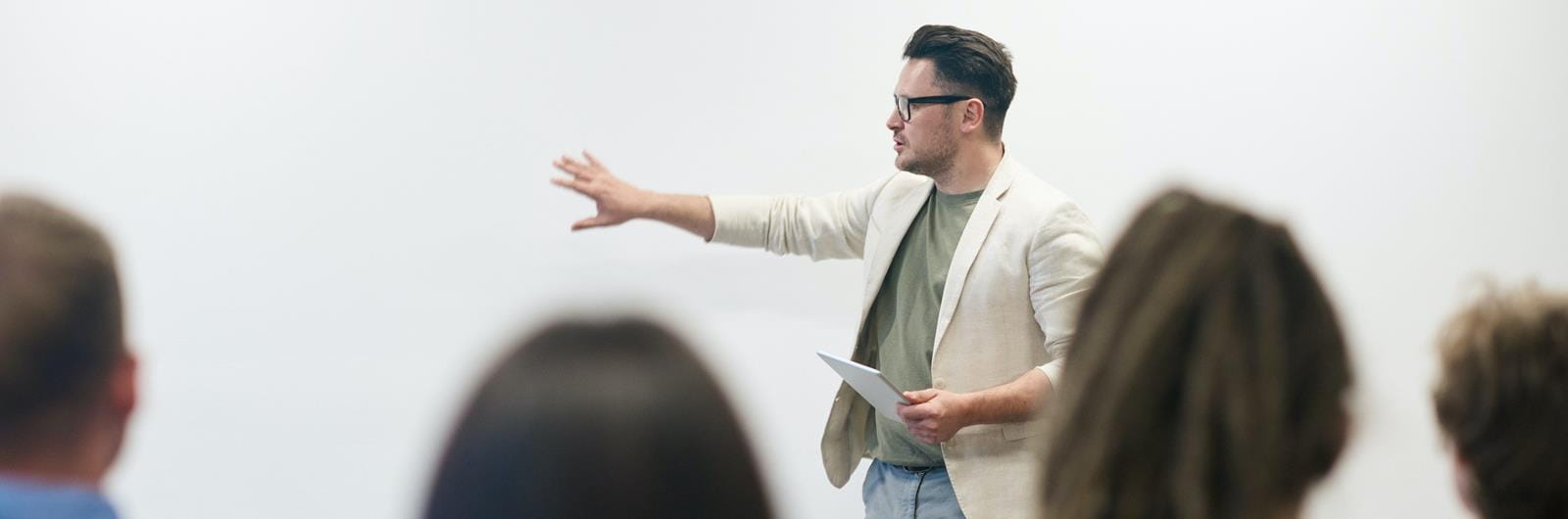 A teacher stands at the front of a room, gesturing while holding a tablet, as students listen attentively.