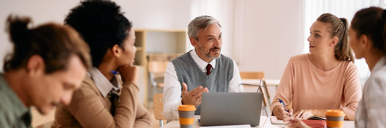 Mature professor uses laptop while talking to group of students in classroom.