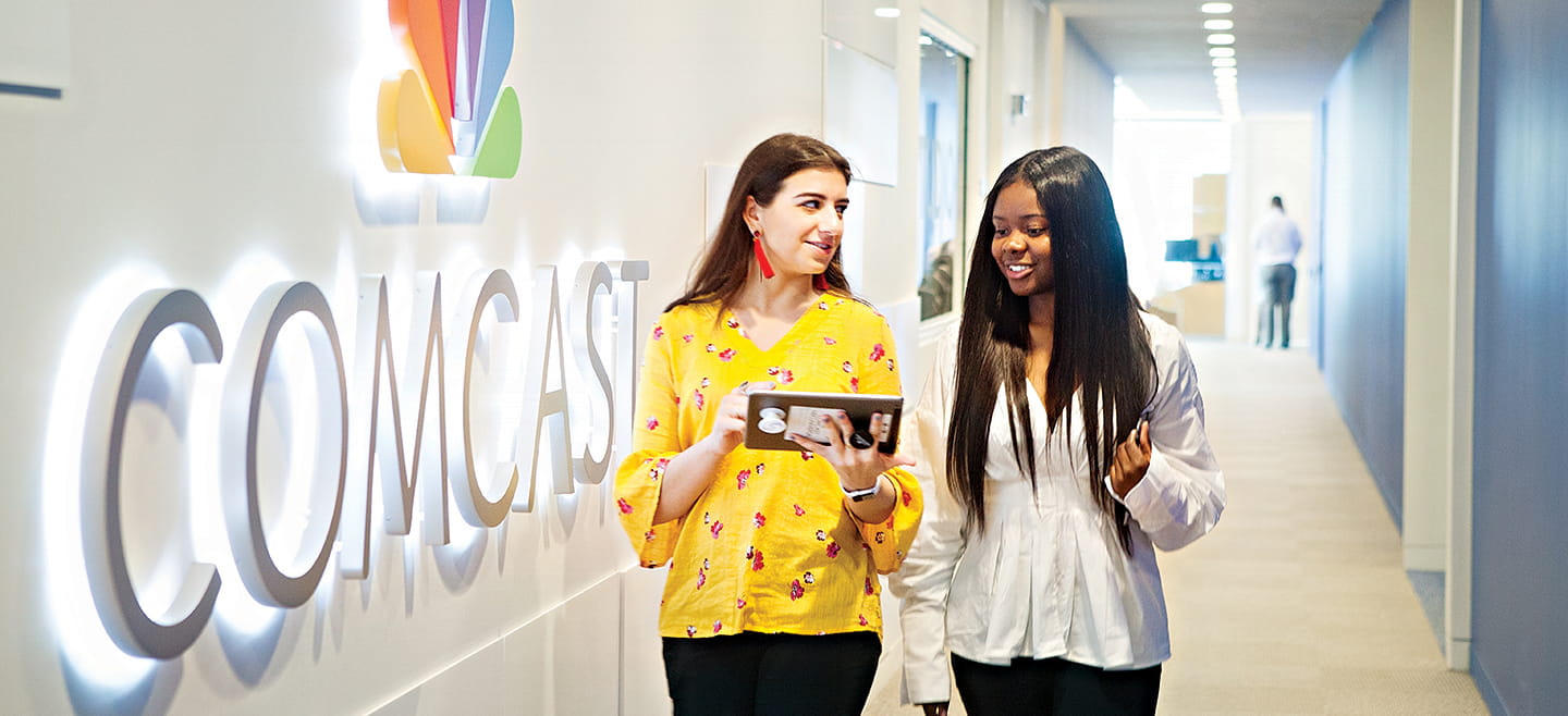 Two women look at a tablet as they walk past an illuminated Comcast sign in a corporate office.