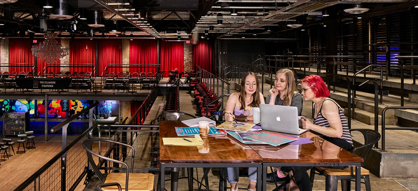 Three women seated at a table overlooking a modern performance space look at a laptop.
