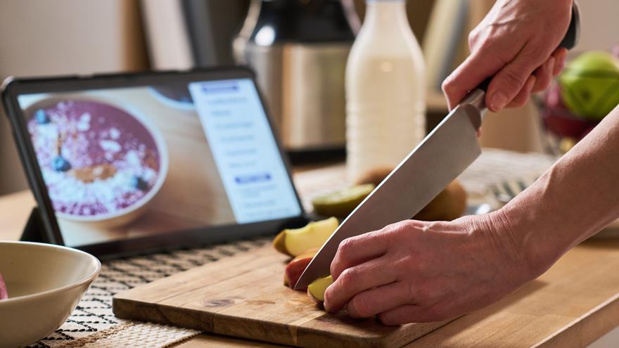 Hands cut an apple with a knife while following a recipe on a tablet. 