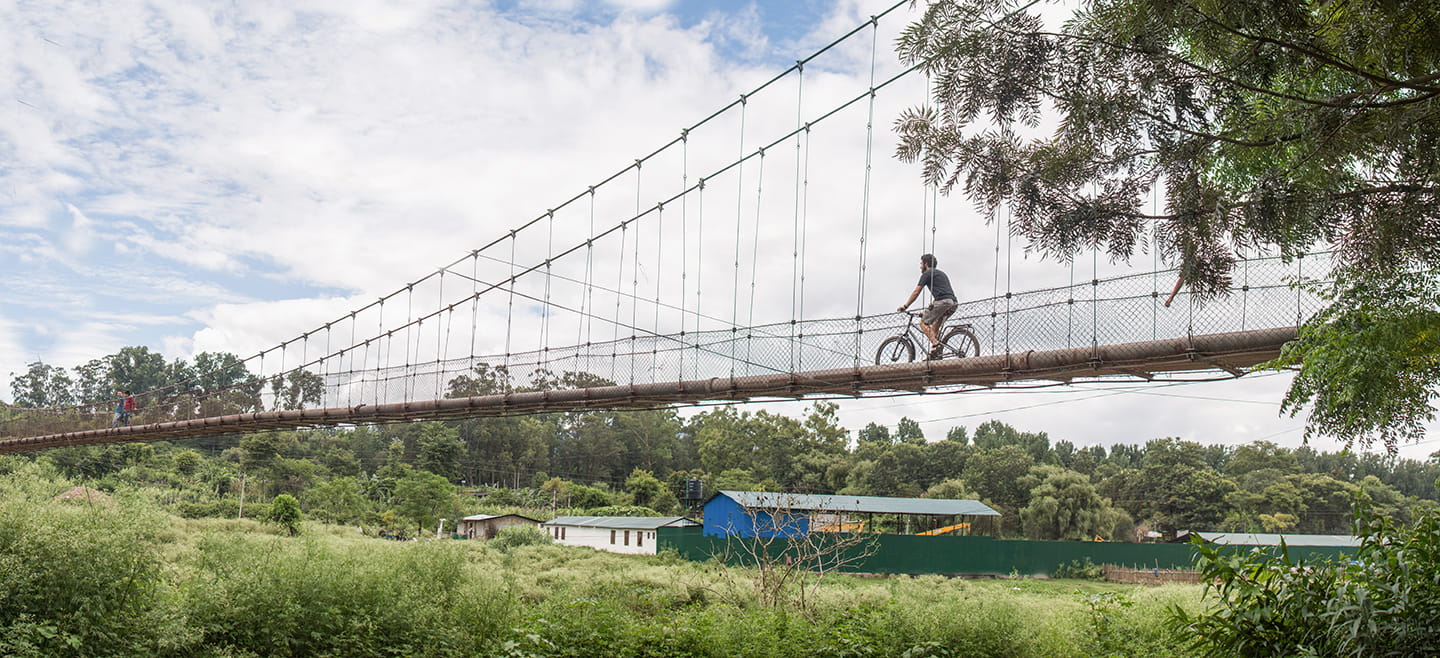 A man rides a bike across a walking bridge suspended over a field.