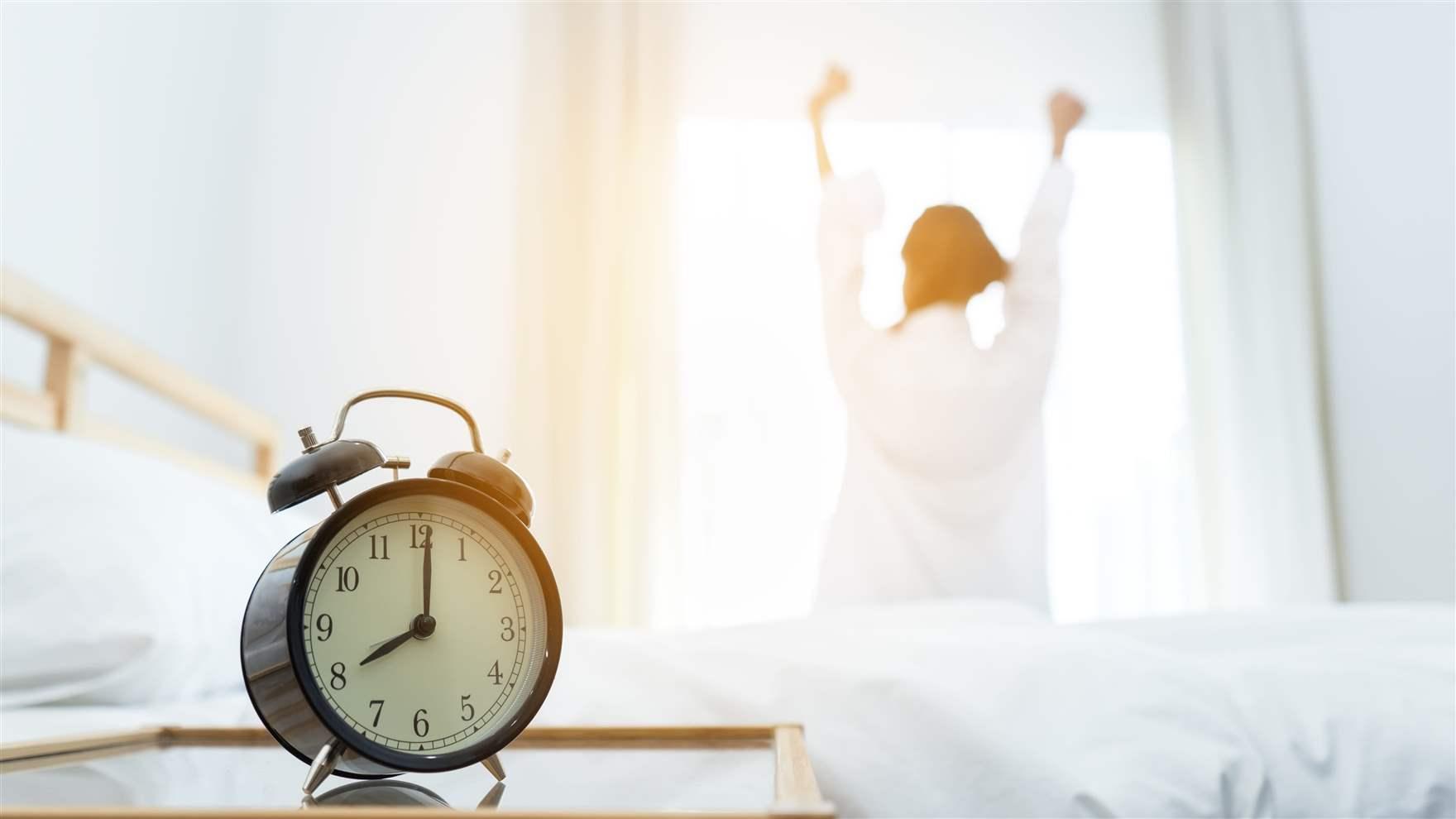 Close-up of an alarm clock as a woman wakes up in the morning and is sitting on the bed at mirror door side relaxing with sunlight.