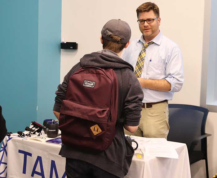 A student wearing a backpack and hat is pictured from behind, talking to a man in a dress shirt and tie who is standing behind a table at a career fair. The tablecloth has the logo for Tamman Inc. printed on it. The man and student are in a conversation. 