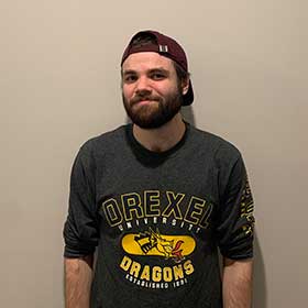 Drexel alumni MJ Rinehart wearing a Drexel Dragons shirt and baseball cap, photographed in front of a white wall.