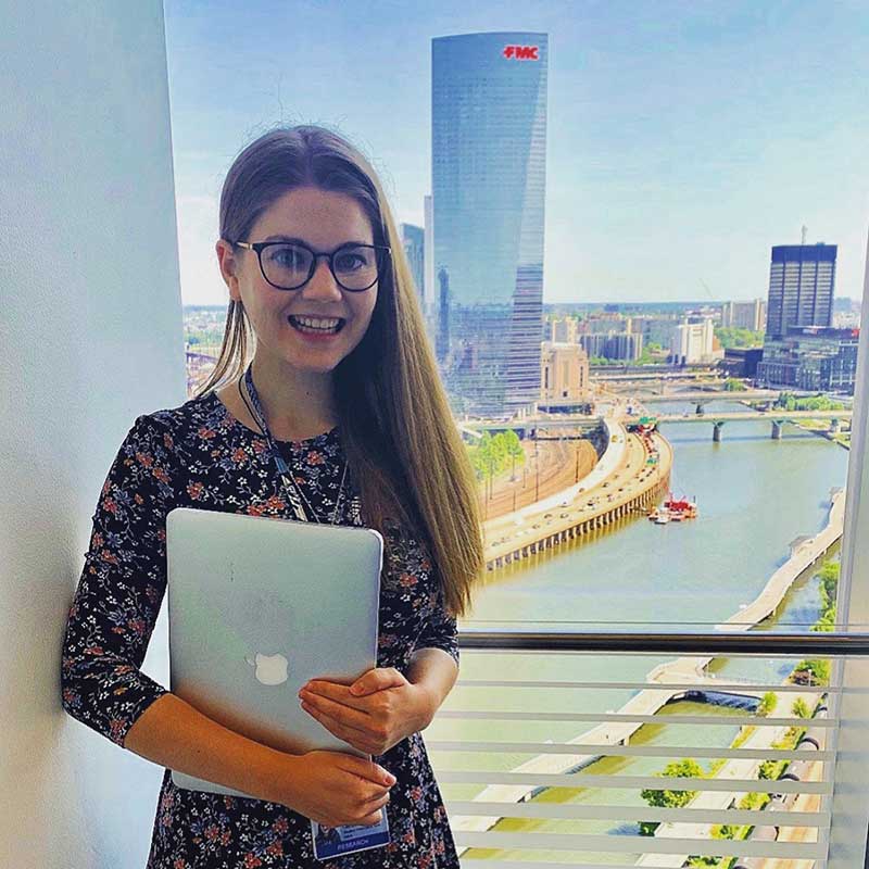 PhD student Elizabeth Campbell photographed in front of a window through which you can see the Philadelphia skyline and FMC building. She is wearing glasses and a floral print dress, holding a laptop and smiling at the camera.
