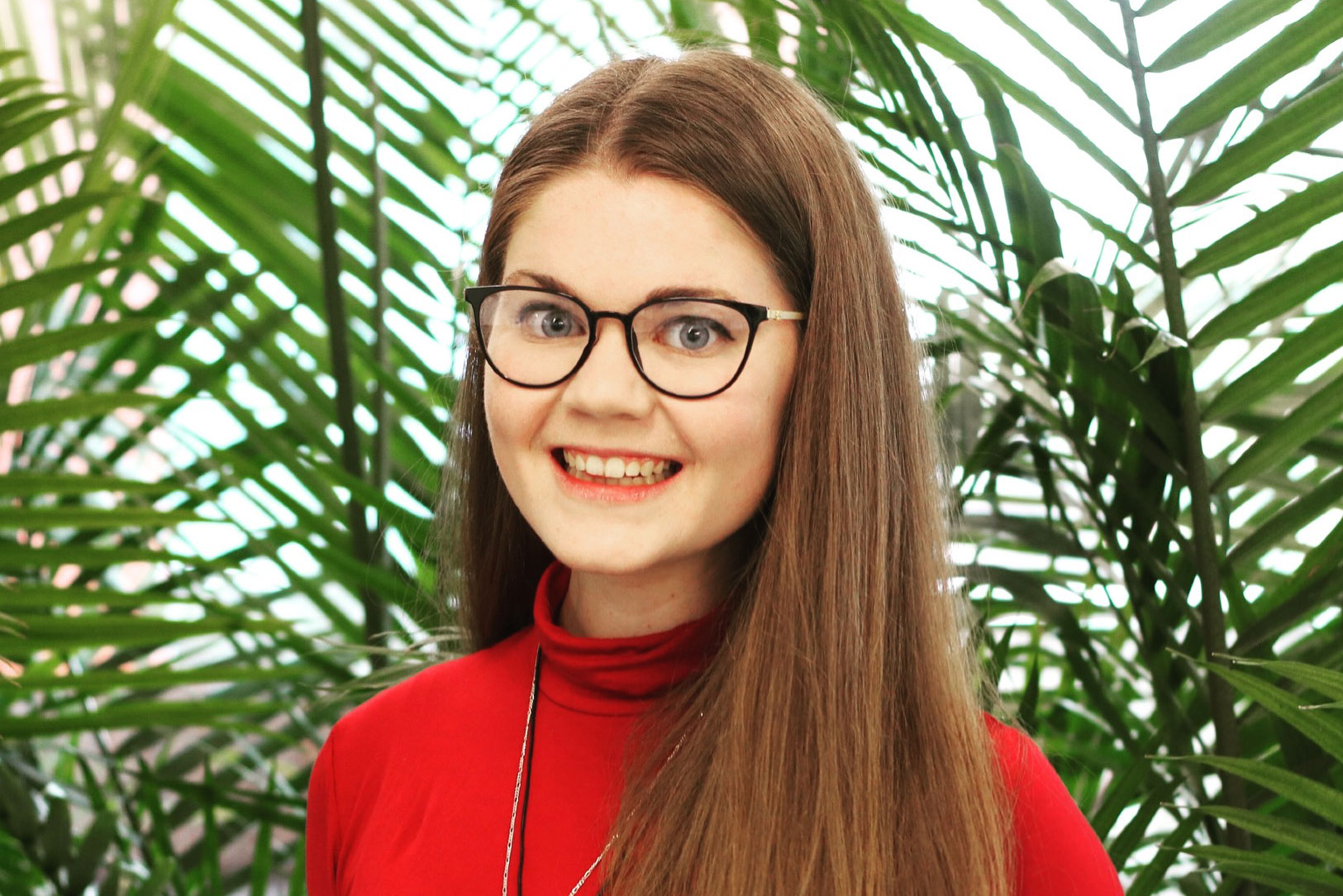 PhD student Elizabeth Campbell photographed from the shoulders up in front of a background of tropical plants. She is wearing a red sweater and glasses, smiling at the camera.